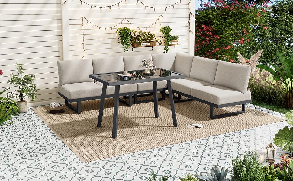 Aménagement de terrasse extérieur comprenant un canapé sectionnel rembourré gris disposé autour d'une table à cadre en métal noir, posé sur des pavés à motifs géométriques avec une pergola blanche recouverte de lierre et de la verdure en arrière-plan.