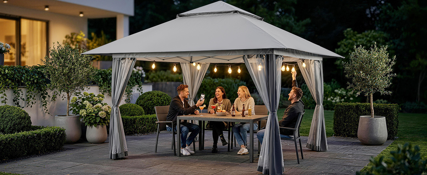 Tente de belvédère hexagonale moderne avec tissu gris et structure en métal située dans la cour du soir. L'intérieur illuminé montre un groupe de personnes assises à une table à manger, entouré de plantes en pot et de meubles de patio sur un sol en pierre.
