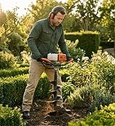 Homme en chemise verte et pantalon beige utilisant une tarière portative ou un semoir dans un jardin avec des haies et une végétation verte en arrière-plan.