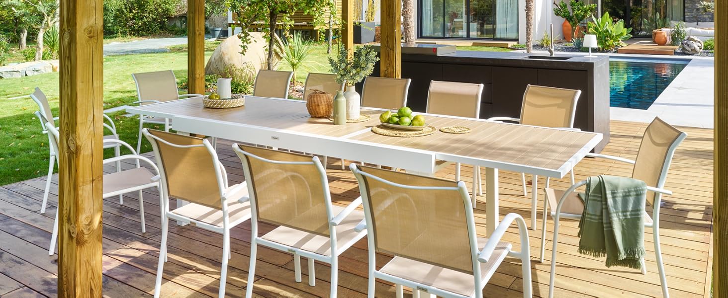 Pergola en bois avec toit de chaume au-dessus de la salle à manger extérieure. Grande table avec chaises blanches sur terrasse en bois. La cuisine extérieure est visible en arrière-plan.