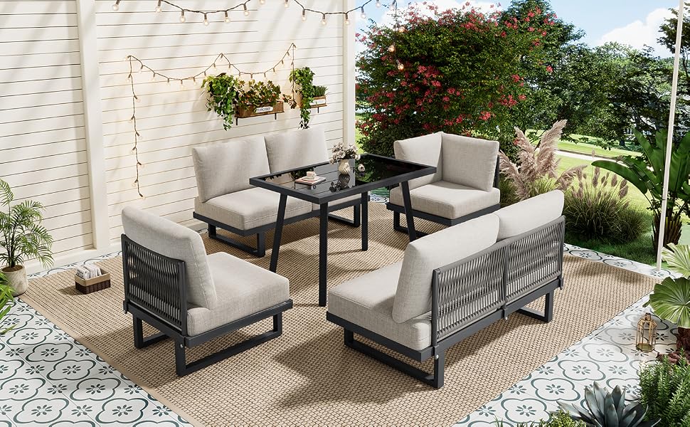 Ensemble de mobilier de conversation pour terrasse extérieure comprenant quatre fauteuils rembourrés gris avec des cadres en métal noir disposés autour d'une table rectangulaire noire sur un tapis d'extérieur à motifs, positionnés sur un patio en brique avec du lierre grimpant et des guirlandes lumineuses.