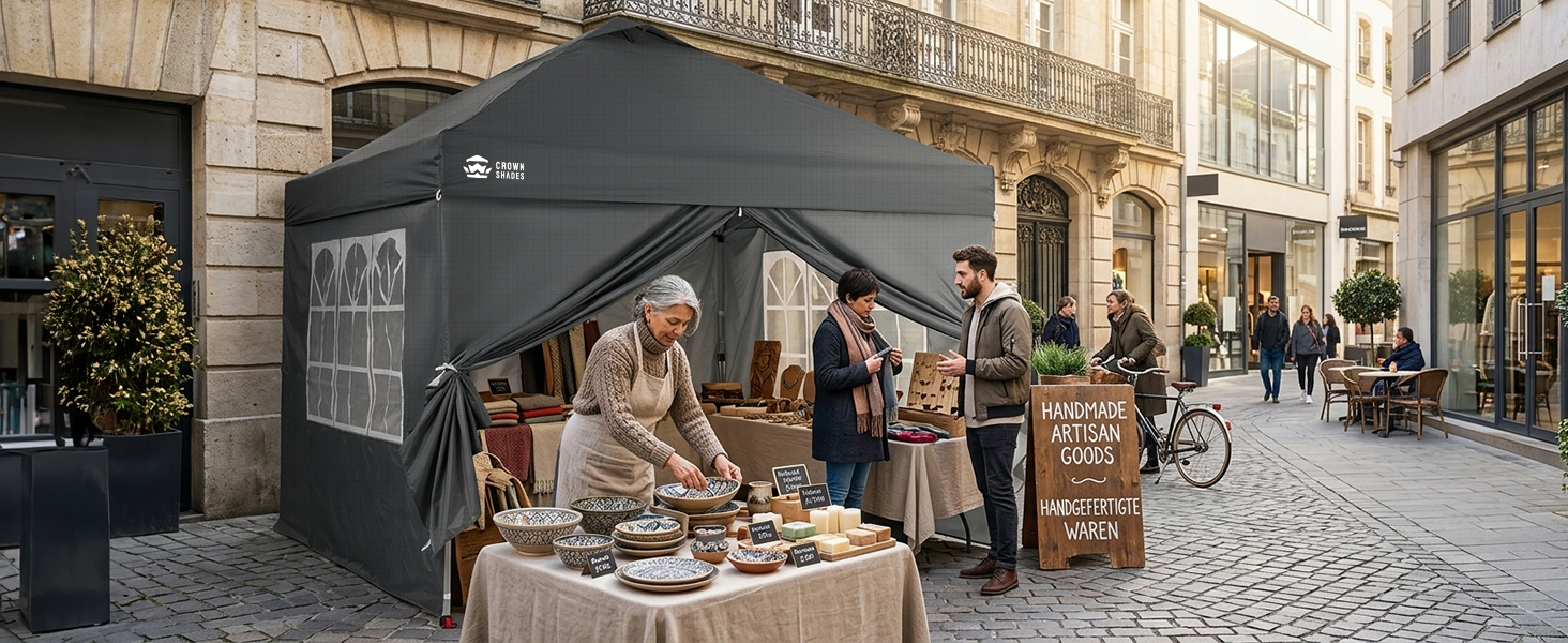 Étal de marché extérieur avec tente à baldaquin noir sur une rue pavée. Le vendeur expose les marchandises sur un comptoir en bois. Bâtiments historiques de style européen et piétons visibles en arrière-plan.