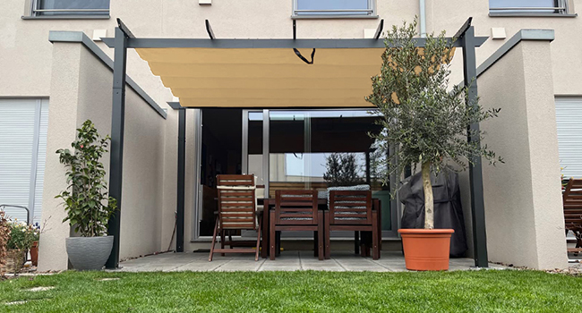 Terrasse extérieure avec auvent beige rétractable soutenu par un cadre en métal noir. Meubles en bois placés en dessous. Plantes en pot et gazon visibles.