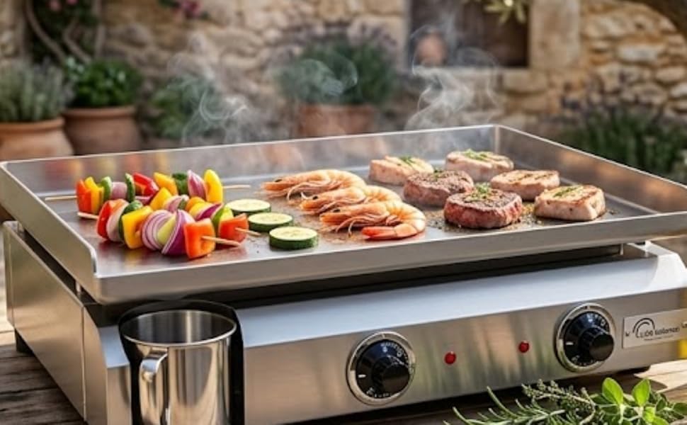Table de cuisson extérieure en acier inoxydable avec deux brûleurs placés sur une table en bois, chargée de légumes et de viandes grillés sur fond de bâtiment en pierre et de jardin.