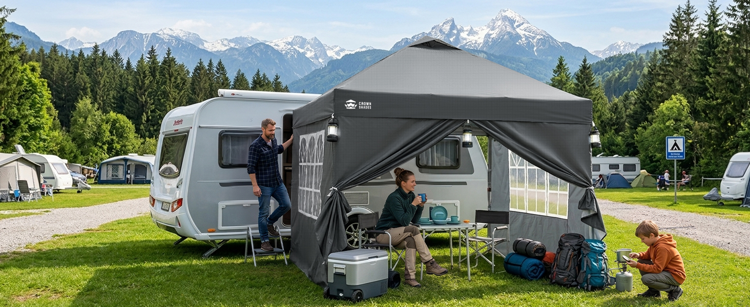 Roulotte de voyage avec toit relevable garée dans un terrain de camping gazonné avec des montagnes en arrière-plan. Une famille de quatre personnes a aménagé un coin salon extérieur avec du matériel de camping à côté du camping-car blanc et gris.