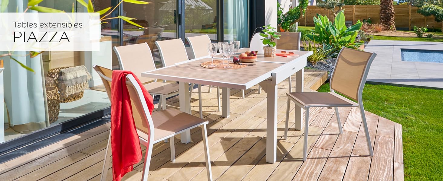 comprenant une grande table en bois et six chaises en maille beige. Serviette rouge drapée sur la chaise. Cadre au bord de la piscine avec plantes et portes vitrées visibles.