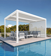 Pergola en aluminium blanc avec toit à persiennes orientables au bord d'une piscine. Mobilier d'extérieur gris posé en dessous sur une terrasse en bois.